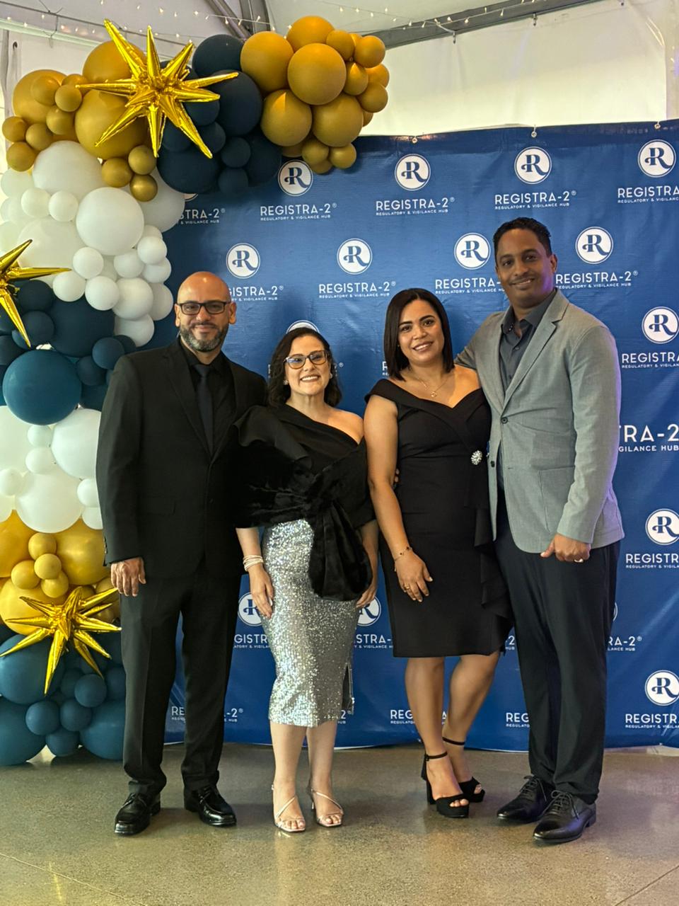 Group of four adults posing for a photo at a formal event with a blue backdrop and balloon arch in gold, white, and teal colors behind them.